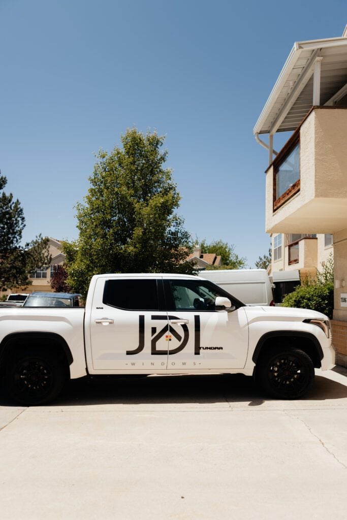 A white truck parked in front of a house with a sign for JDI Windows visible in the yard.