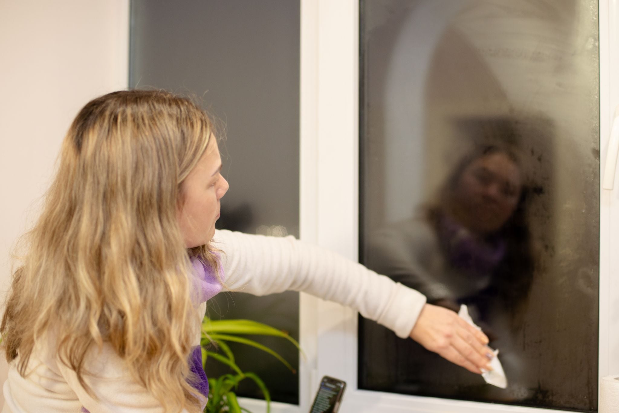 A blonde woman wiping condensation off a window pane with a cloth.