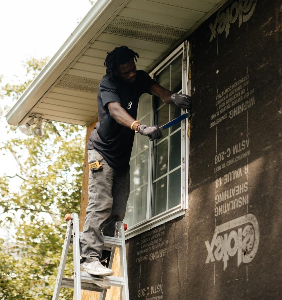 A man painting the exterior of a house using a brush and ladder with trees and a clear sky in the background