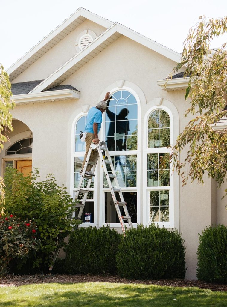 A man on a ladder cleaning the windows of a house wearing safety gear and focused on his task
